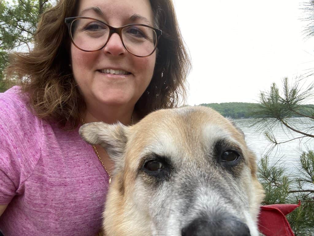 The author is wearing a pink shirt, and smiling into the camera with her dog, Lucy in the foreground. There is a lake in the background.